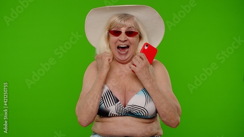 Mature woman traveler in sunglasses browsing on mobile phone and celebrating winning holiday resort vacation. Senior grandmother in swimsuit bra on chroma key background. Tourism, summer trip to sea