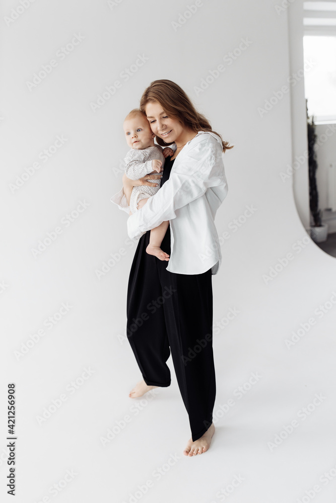 Stylish young mother holding her adorable baby girl in arms, smiling. Attractive beautiful woman gently hugging cute little daughter. Photo in the studio on a white background
