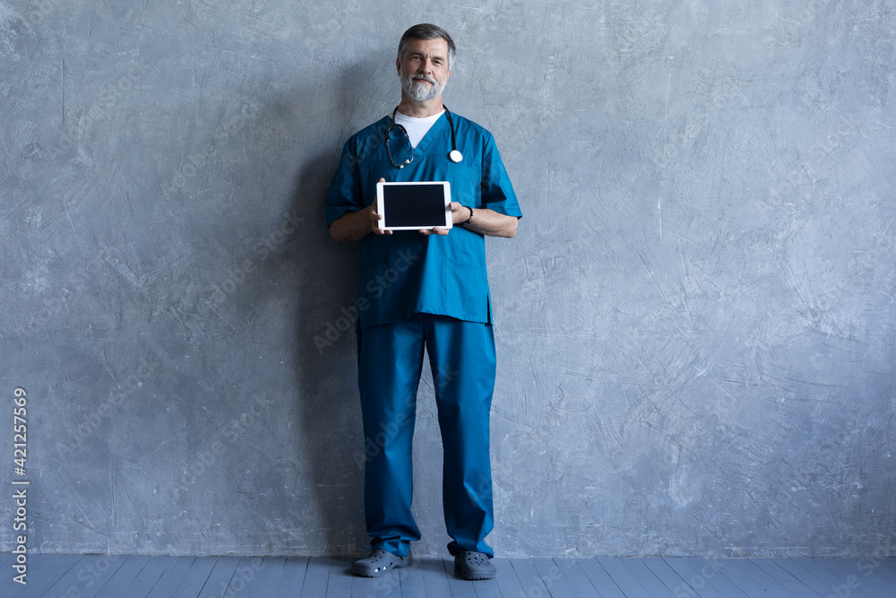 Portrait of professional mature surgeon holding a tablet, looking at camera while standing against the grey background. Portrait of professional mature surgeon holding a tablet, looking at camera while standing against the grey background.