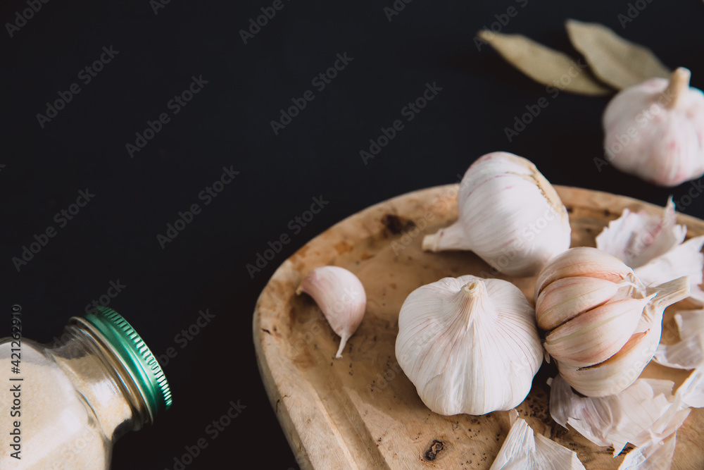 Garlic bulbs on wooden board on  black background, with dried garlic and bay leaves. Condiments and spices in kitchen.