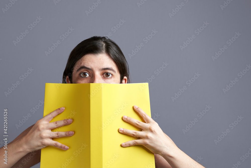 Young woman looking at camera, peeking out from behind an open yellow book Young woman looking at camera, peeking out from behind an open yellow book