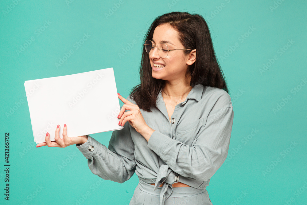 Photo of cute female in glasses holds papers. Wears grey shirt, isolated turquoise color background Photo of cute female in glasses holds papers. Wears grey shirt, isolated turquoise color background