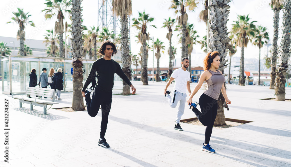 Obraz premium Attractive female fitness instructor in electronic headphones showing exercising method to ethletic clients enjoying morning workout, group of diverse people collaborating together on stretching