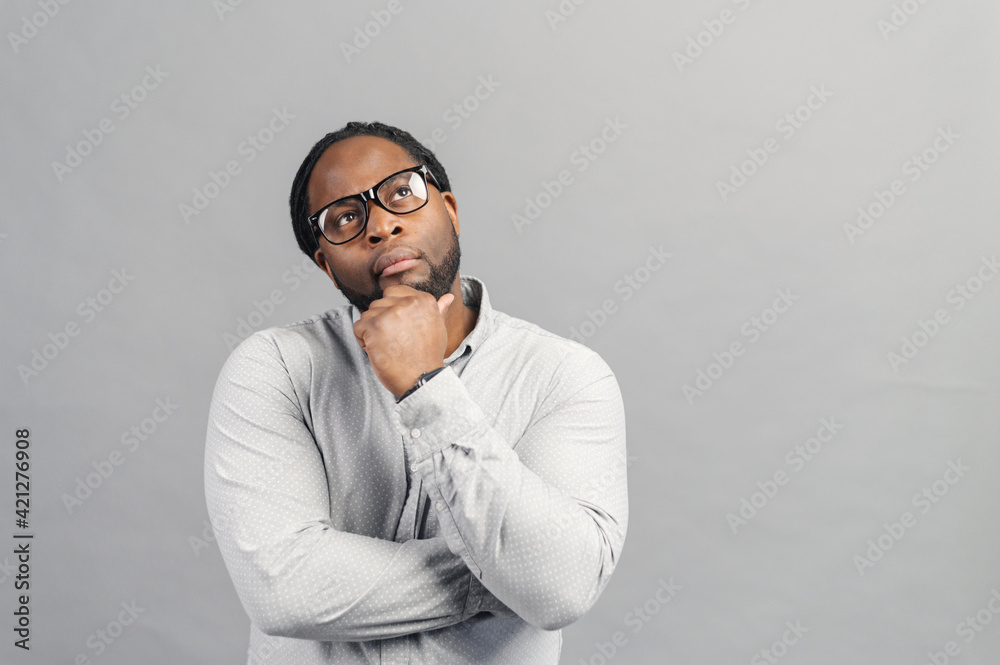 Fototapeta premium Thoughtful African-American man wearing stylish eyeglasses and smart casual shirt looking new ideas and inspiration, holds chin with a head , looks up, lost in thoughts, isolated on grey