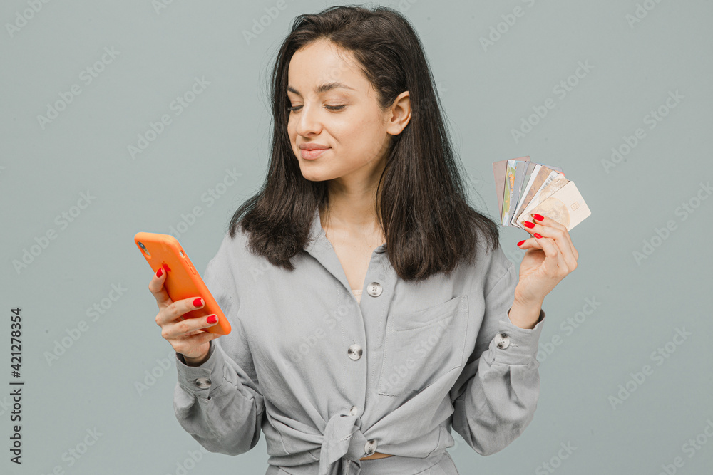 Photo of cute smiling female holds smartphone and credit cads. Wears grey shirt, isolated grey color background Photo of cute smiling female holds smartphone and credit cads. Wears grey shirt, isolated grey color background