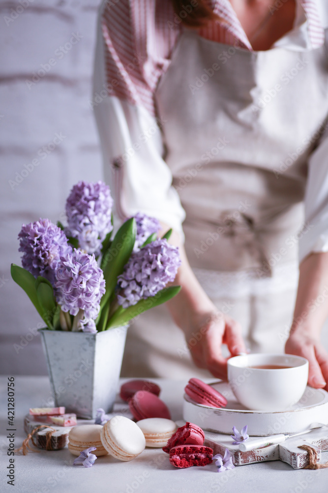 Spring atmosphere. Flowers in a vase, macaroni, chocolate and a cup of tea on wooden white boards on a light table with hands. Lilac hyacinths. Background image, copy space, vertical, rustic
