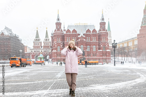 A beautiful young girl in a pink jacket walks along Manezhnaya Square in Moscow during a snowfall and blizzard. Snowblowers are working in the background.