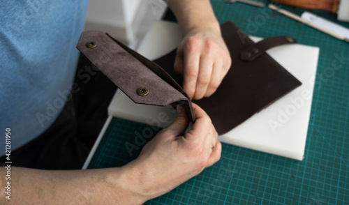 Mens hand working on the leather wallet in his workshop. Working process with a brown natural leather. Craftsman holding a crafting tools.