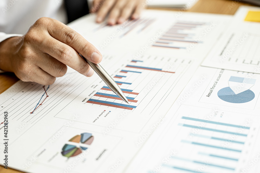 A close-up of a financial businessman holding a pen and pointing at the information sheet on his desk, reading the company's financials to make a financial plan. Financial concept. A close-up of a financial businessman holding a pen and pointing at the information sheet on his desk, reading the company's financials to make a financial plan. Financial concept.