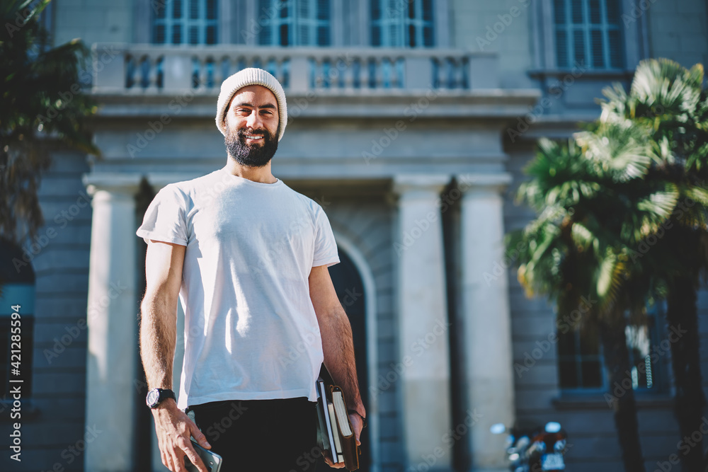 Half length portrait of cheerful Turkish student in hat smiling at camera holding education equipment in hand, happy Middle Eastern male with textbooks posing at street urban setting on leisure - obrazy, fototapety, plakaty 