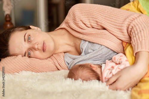 Soft photo of young mother feeding breast her baby at home while lying in bed  mommy looking at camera with tired and sleepless facial expression  wearing home clothing.