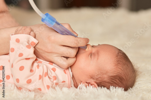 Slika na platnu Faceless mother using nasal aspiration sucks the snot from baby, child lying on bed on fluffy blanket with closed eyes, unknown person taking care of infant