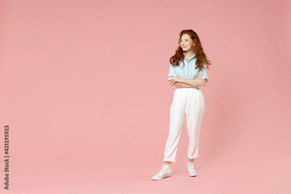 Full length body young student positive smiling happy caucasian european redhead woman 20s in blue shirt pants hold hands crossed folded look aside isolated on pastel pink background studio portrait. Full length body young student positive smiling happy caucasian european redhead woman 20s in blue shirt pants hold hands crossed folded look aside isolated on pastel pink background studio portrait.