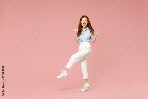Full length body fun young student lucky overjoyed excited happy redhead woman 20s in blue shirt pants walk do winner gesture clench fist celebrate isolated on pastel pink background studio portrait