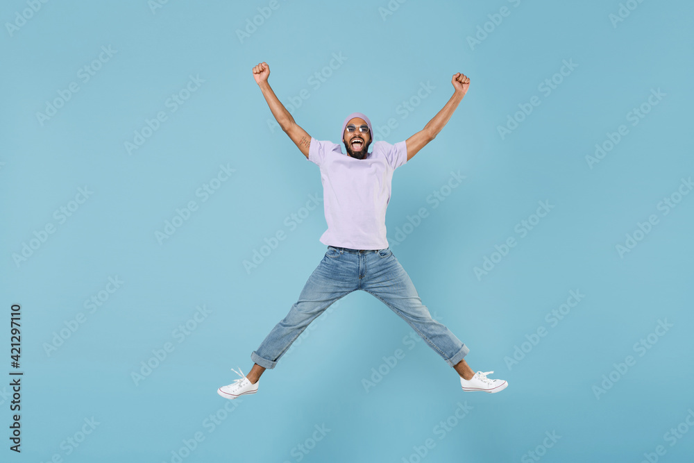 Full length young overjoyed excited happy cheerful unshaven black african man in violet t-shirt hat glasses jump high with outstretched legs hands isolated on pastel blue background studio portrait.