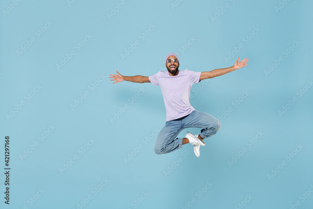 Full length young overjoyed excited cool happy cheerful unshaven black african man in violet t-shirt hat glasses jump high with outstretched hands isolated on pastel blue background studio portrait