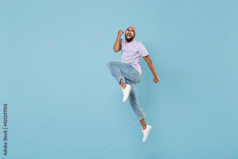 Full length young overjoyed unshaven black african man in violet t-shirt hat glasses do winner gesture jump high clench fist celebrate scream yes isolated on pastel blue background studio portrait Full length young overjoyed unshaven black african man in violet t-shirt hat glasses do winner gesture jump high clench fist celebrate scream yes isolated on pastel blue background studio portrait