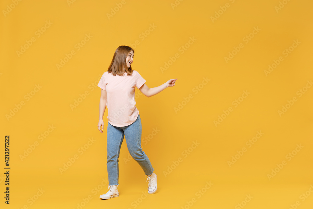 Full length of young caucasian woman 20s wearing casual basic pastel pink t-shirt, jeans pointing index finger aside on workspace area mock up copy space isolated on yellow background studio portrait. Full length of young caucasian woman 20s wearing casual basic pastel pink t-shirt, jeans pointing index finger aside on workspace area mock up copy space isolated on yellow background studio portrait.