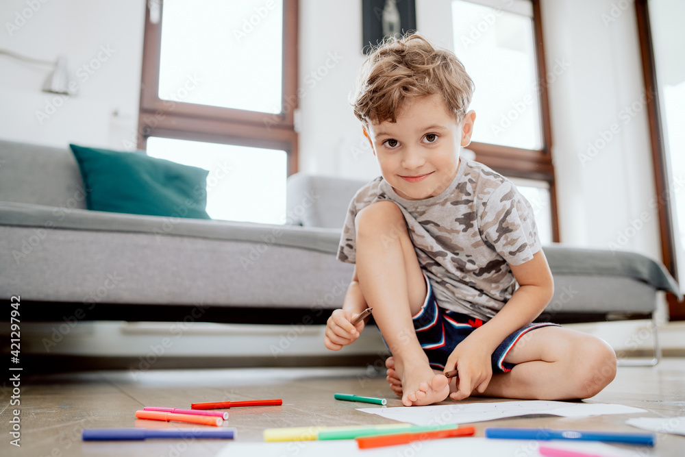 Portrait of a school-aged boy sitting on a wooden floor in the living room of a house. The boy holds a marker in his hand and looks into the camera. Portrait of a school-aged boy sitting on a wooden floor in the living room of a house. The boy holds a marker in his hand and looks into the camera.