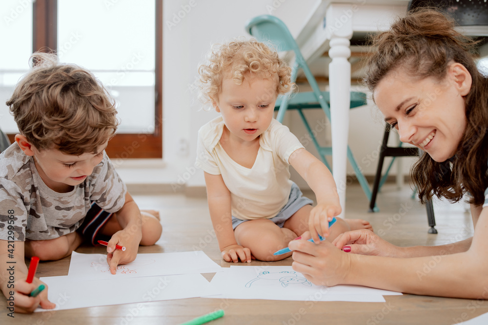 Portrait of Mom with her school-aged son and preschool-aged daughter drawing together while sitting on a wooden floor in the living room at home.