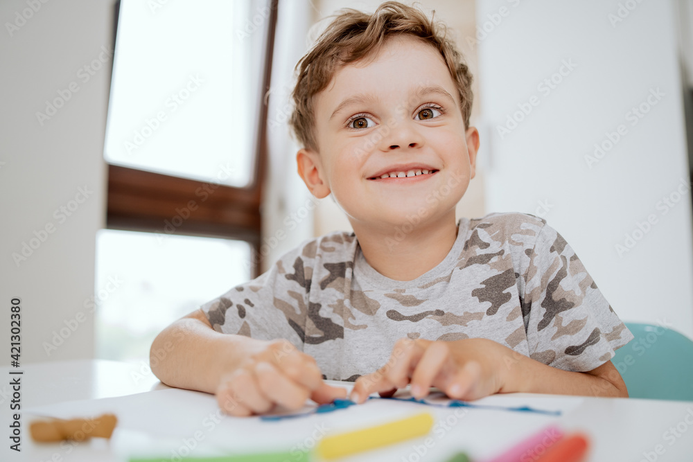 Portrait of a lovely happy school-aged boy sitting at kitchen table enjoying some free time. Little boy is making something out of plasticine. Portrait of a lovely happy school-aged boy sitting at kitchen table enjoying some free time. Little boy is making something out of plasticine.
