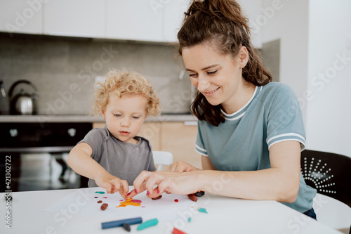 A beautiful mommy sits at the kitchen table with her preschool-aged daughter and makes something out of plasticine. Mom is spending her afternoon free time with her daughter.