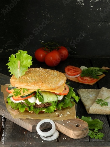 sandwich with ham and vegetables. Burger with sesame bun, cucumber, tomato, cheese, onion, sauce and salad, lies on a dark cutting board on a black background. Photo for the recipe.Tomatoes lie behind photo