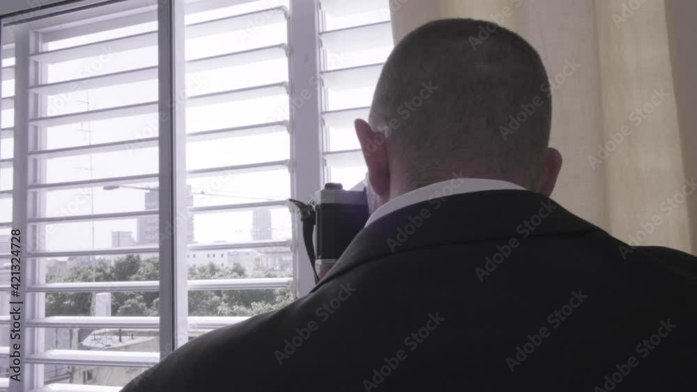 Man taking pictures from a window. Over the shoulder of a guy holding a black camera and filming people from a window with shutters. Undercover detective spying and paparazziing from inside his house. Man taking pictures from a window. Over the shoulder of a guy holding a black camera and filming people from a window with shutters. Undercover detective spying and paparazziing from inside his house.