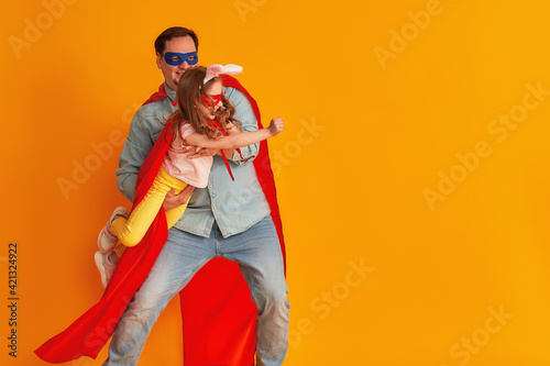 serious father with a beard, a superhero holding a little baby girl, together dressed in superman costumes, preparing for a children's party, isolated on a yellow wall. Strong heroes. copy-space