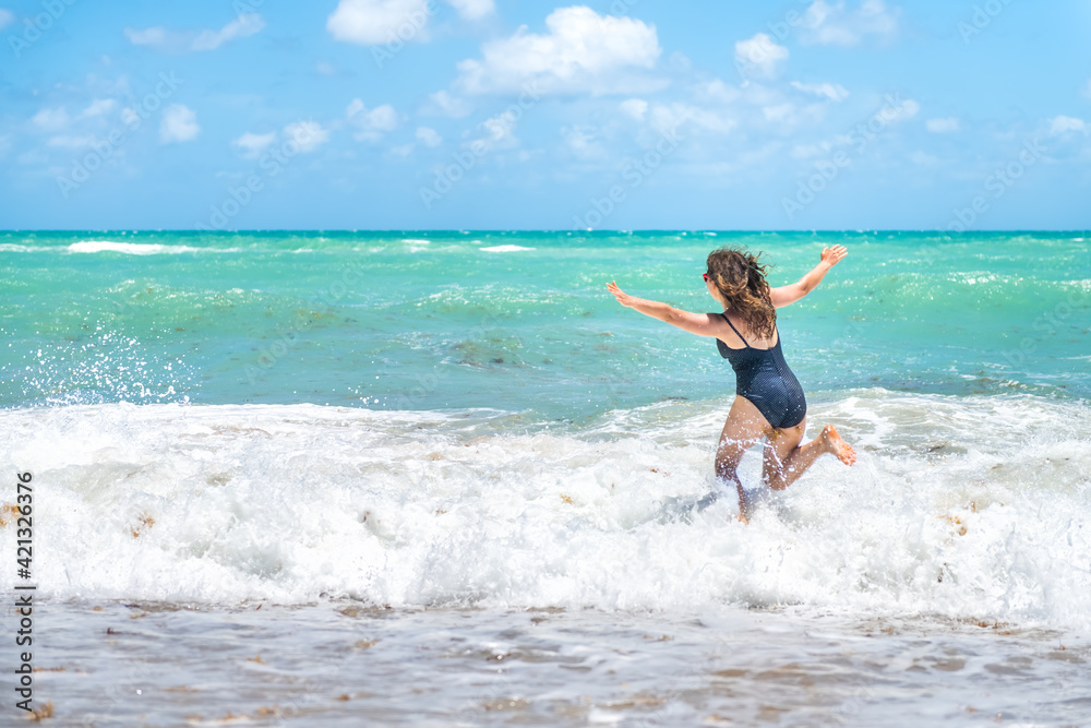 Back of young woman running in polka dot swimsuit with waves splashing crashing in Hollywood, Miami Beach Florida with Atlantic ocean sea green water in summer Back of young woman running in polka dot swimsuit with waves splashing crashing in Hollywood, Miami Beach Florida with Atlantic ocean sea green water in summer