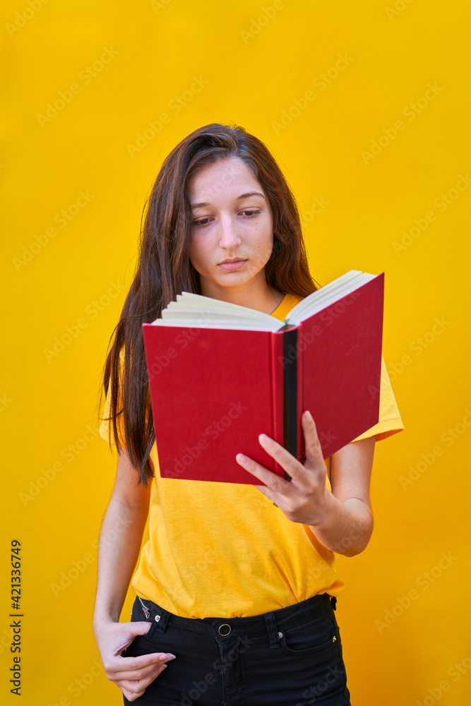 American flat vertical photo of a Caucasian young girl with long brunette hair reading a red book on yellow background American flat vertical photo of a Caucasian young girl with long brunette hair reading a red book on yellow background