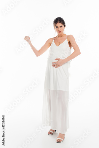 full length view of angry, pregnant bride touching belly and showing clenched fist on white