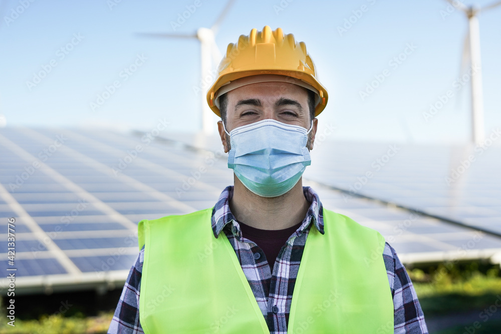 Young man working for renewable energy while wearing safety face mask for coronavirus outbreak - Solar panel station and wind turbines - obrazy, fototapety, plakaty 