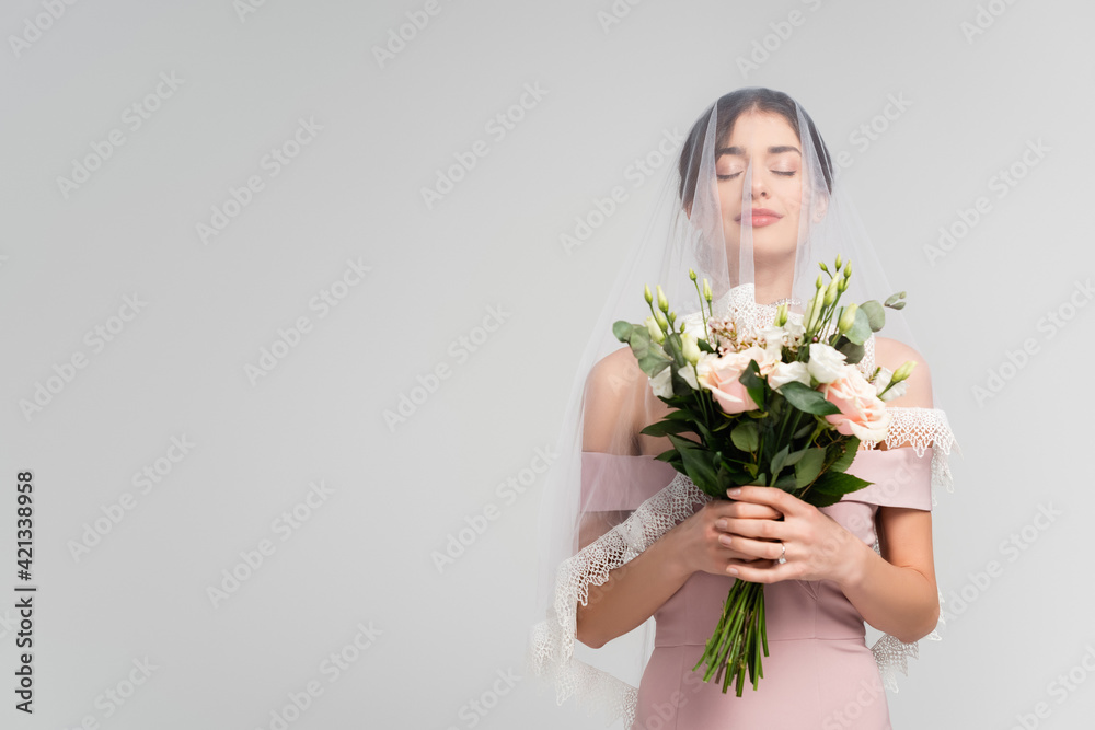 young bride in veil holding wedding bouquet while posing with closed eyes isolated on grey young bride in veil holding wedding bouquet while posing with closed eyes isolated on grey