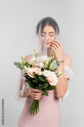 young bride in veil holding wedding bouquet while standing with closed eyes isolated on grey