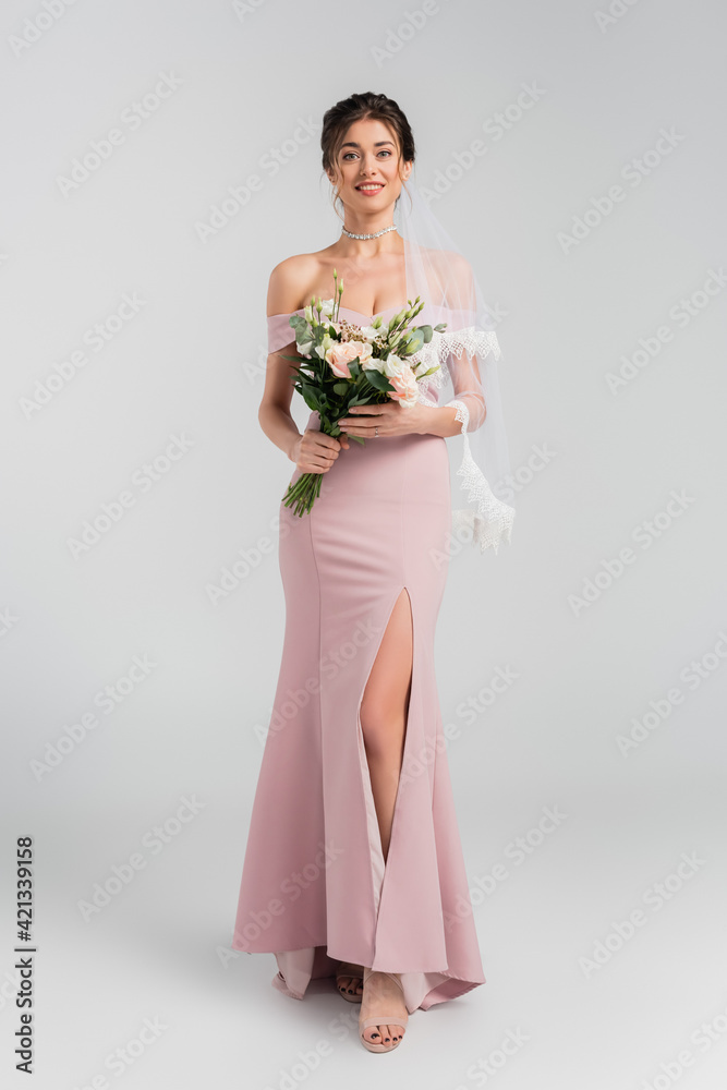 full length view of smiling fiancee in pink dress posing with bouquet on grey full length view of smiling fiancee in pink dress posing with bouquet on grey
