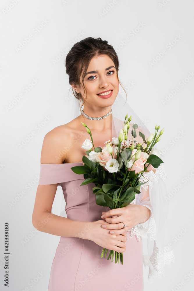 pretty woman in pink dress and veil holding wedding bouquet while smiling at camera isolated on grey pretty woman in pink dress and veil holding wedding bouquet while smiling at camera isolated on grey