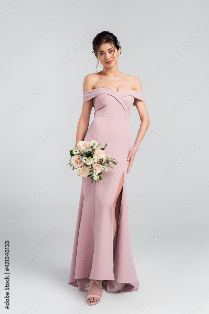 full length view of elegant woman with wedding bouquet looking at camera on grey full length view of elegant woman with wedding bouquet looking at camera on grey