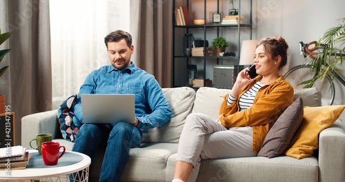 Young married couple resting on sofa in living room using digital gadget. Caucasian handsome man working on laptop browsing online while his pretty wife talking on smartphone. Leisure concept