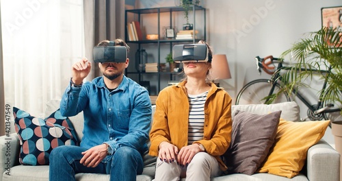 Young Caucasian couple man and woman sitting on couch in room wearing VR glasses using innovative futuristic technologies spending time at home making gesture with hands in air, virtual reality