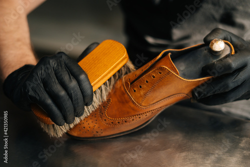 Close-up hands of unrecognizable shoemaker cleaning with brush old light brown leather shoe. Concept of cobbler artisan repairing and restoration work in shoe repair shop.