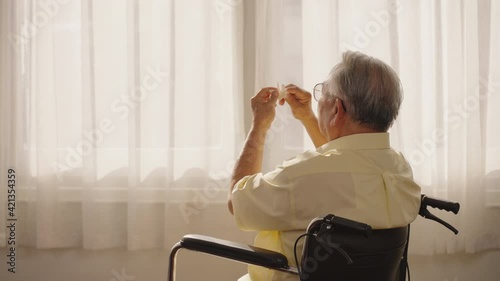 Asian male patient looking outside the window while sitting in wheelchair in living room at retirement home. Asian Thai elderly person sitting lonely in a wheelchair holding a paper bird to play.