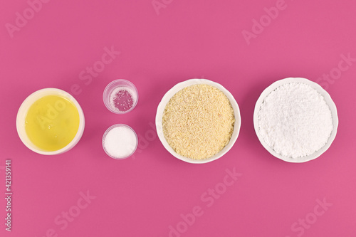 Bowls with ingredients for making homemade French Macaron sweets showing powdered sugar, ground almonds, egg white, salt and sugar on pink background