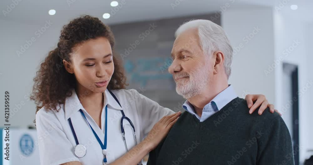 Portrait of cute female multiethnic doctor hugs shoulders senior male patient and congratulates him with health recovery, talk about good news smiling each other standing in hospital