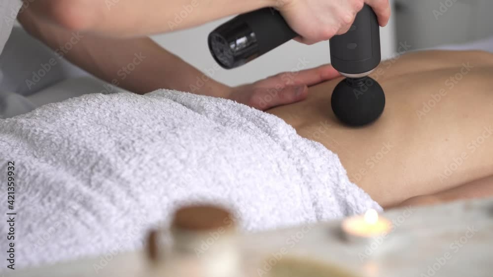 The body massage makes the masseur a percussion massage therapist. The girl lies on her stomach, the masseur does a back massage with a percussion massager. Close-up in a massage parlor. Couch white