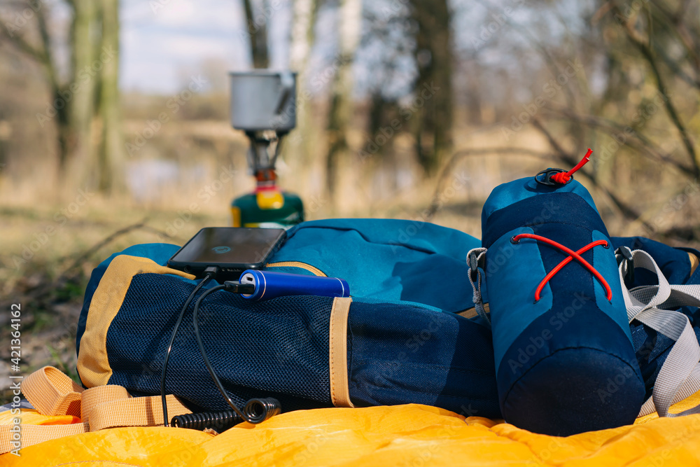 Charge your smartphone while traveling with a power bank. A portable charger charges the phone against the backdrop of a tourist gas burner and a forest. Charge your smartphone while traveling with a power bank. A portable charger charges the phone against the backdrop of a tourist gas burner and a forest.