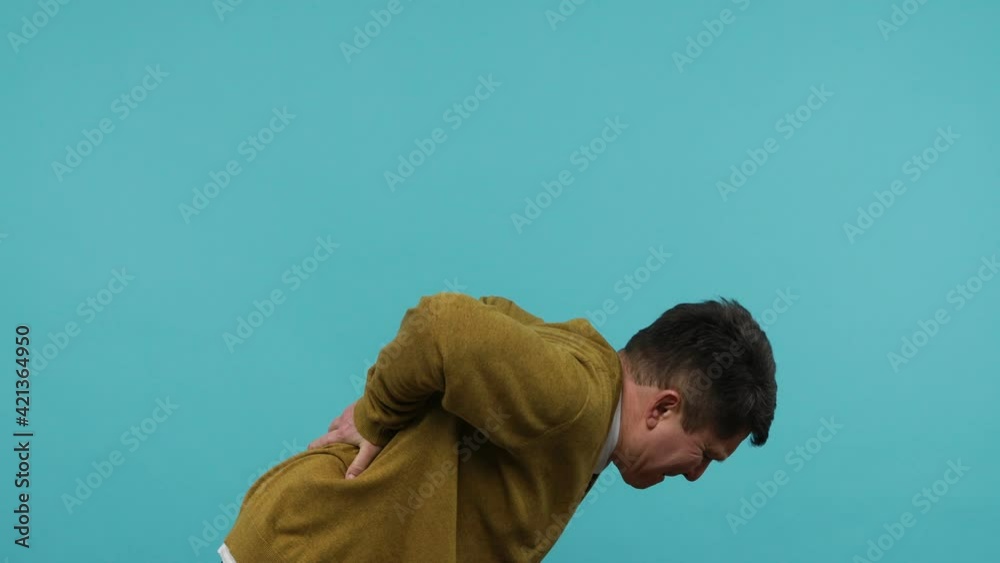 Backache. Unhealthy middle aged dark haired man touching injured sore back, suffering lower lumbar discomfort, muscle pain of overwork, pinched nerve. Indoor studio shot isolated on blue background Backache. Unhealthy middle aged dark haired man touching injured sore back, suffering lower lumbar discomfort, muscle pain of overwork, pinched nerve. Indoor studio shot isolated on blue background