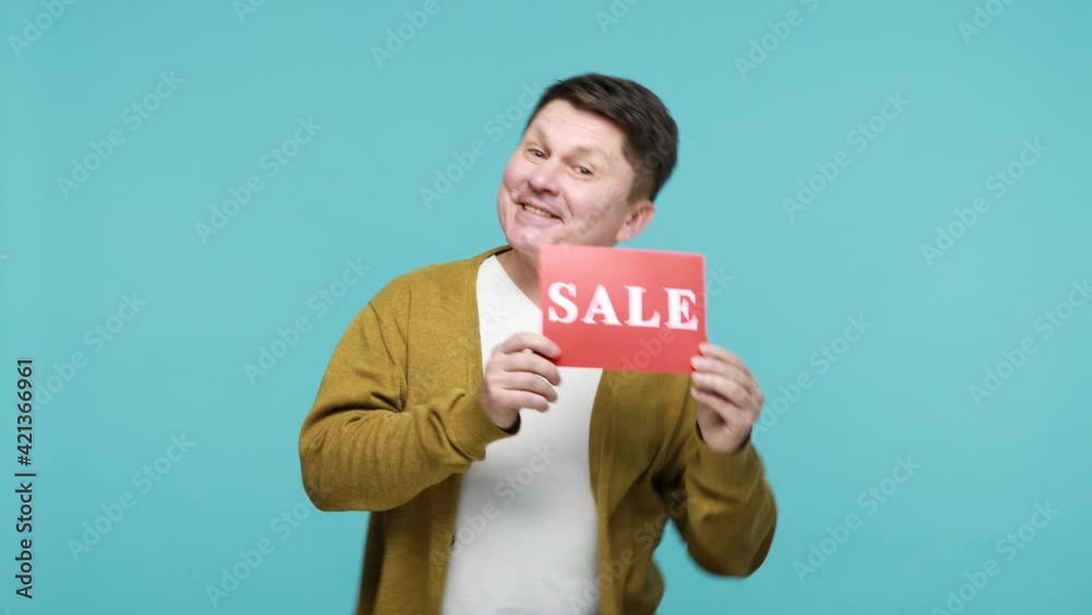 Delighted joyful mature man in white t-shirt and cardigan dancing holding in hands red template with sale inscription, happy satisfied with discounts. Indoor studio shot isolated on blue background
