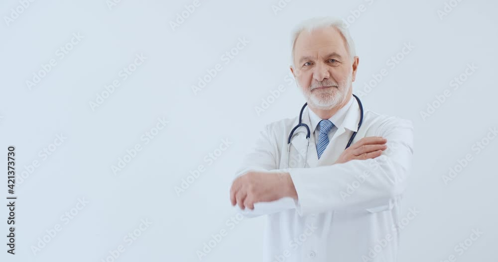 Portrait of senior caucasian male doctor in medical coat crossing arms looking at camera standing on white background in hospital. Handsome therapist in uniform. Healthcare and medicine concept Portrait of senior caucasian male doctor in medical coat crossing arms looking at camera standing on white background in hospital. Handsome therapist in uniform. Healthcare and medicine concept