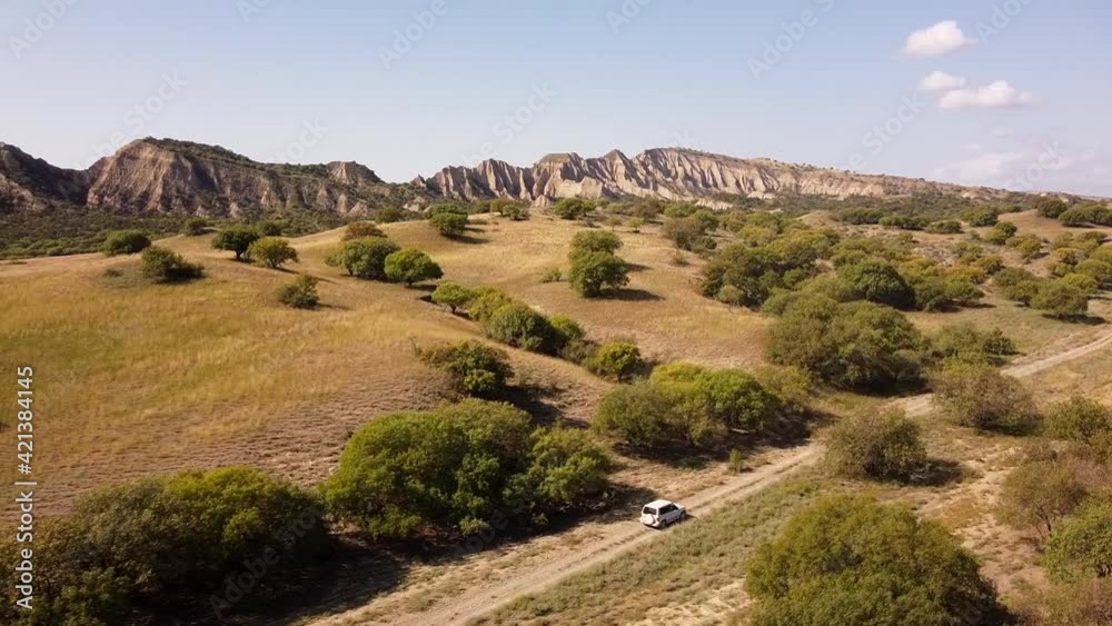 Aerial drone tracking shot of an offroad car driving on a dirt road in the middle of a desert, mountains behind, in Vashlovani National Park, Georgia